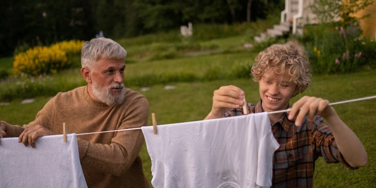 Elderly man and teenage boy joyfully hanging laundry in a sunny garden setting.