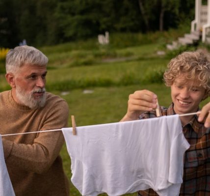 Elderly man and teenage boy joyfully hanging laundry in a sunny garden setting.